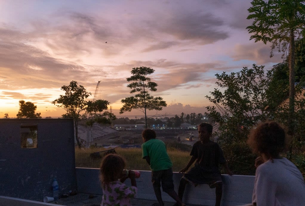 Children sit on a wall at sunset overlooking a construction site in the distance.