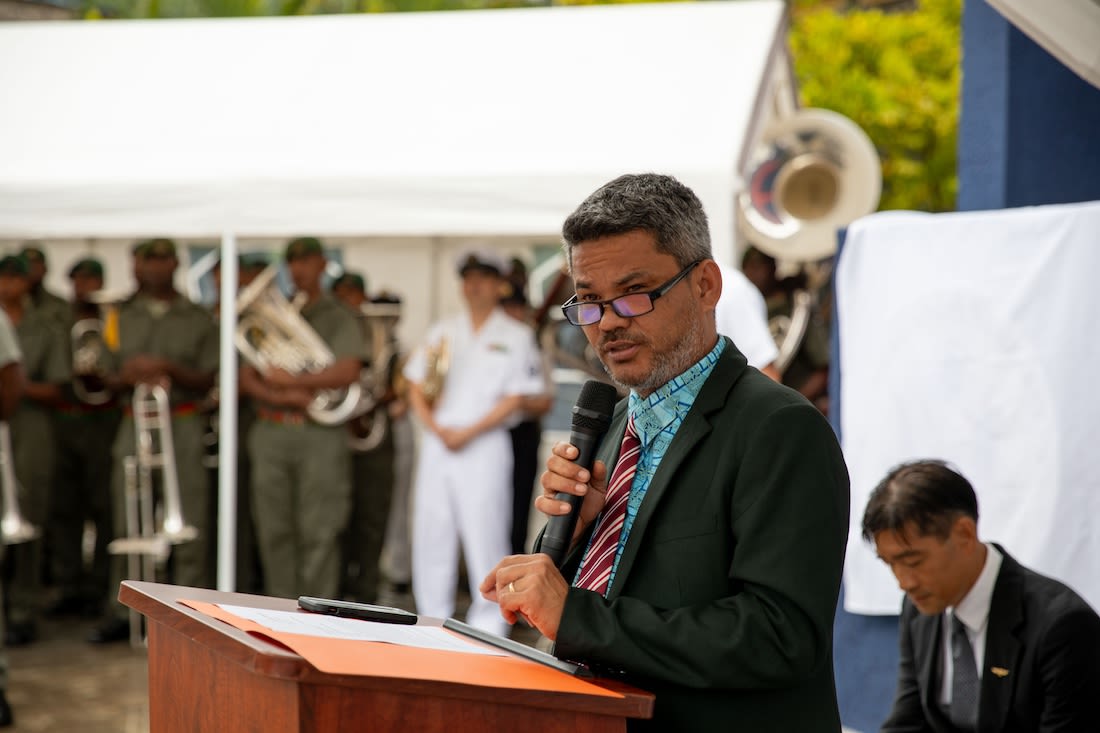 A man makes a speech at a podium as men in uniform holding band instruments stand in the background.