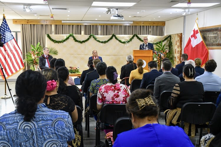 People sit in a room with the U.S. and Tongan flags as leaders wearing leis make speeches.