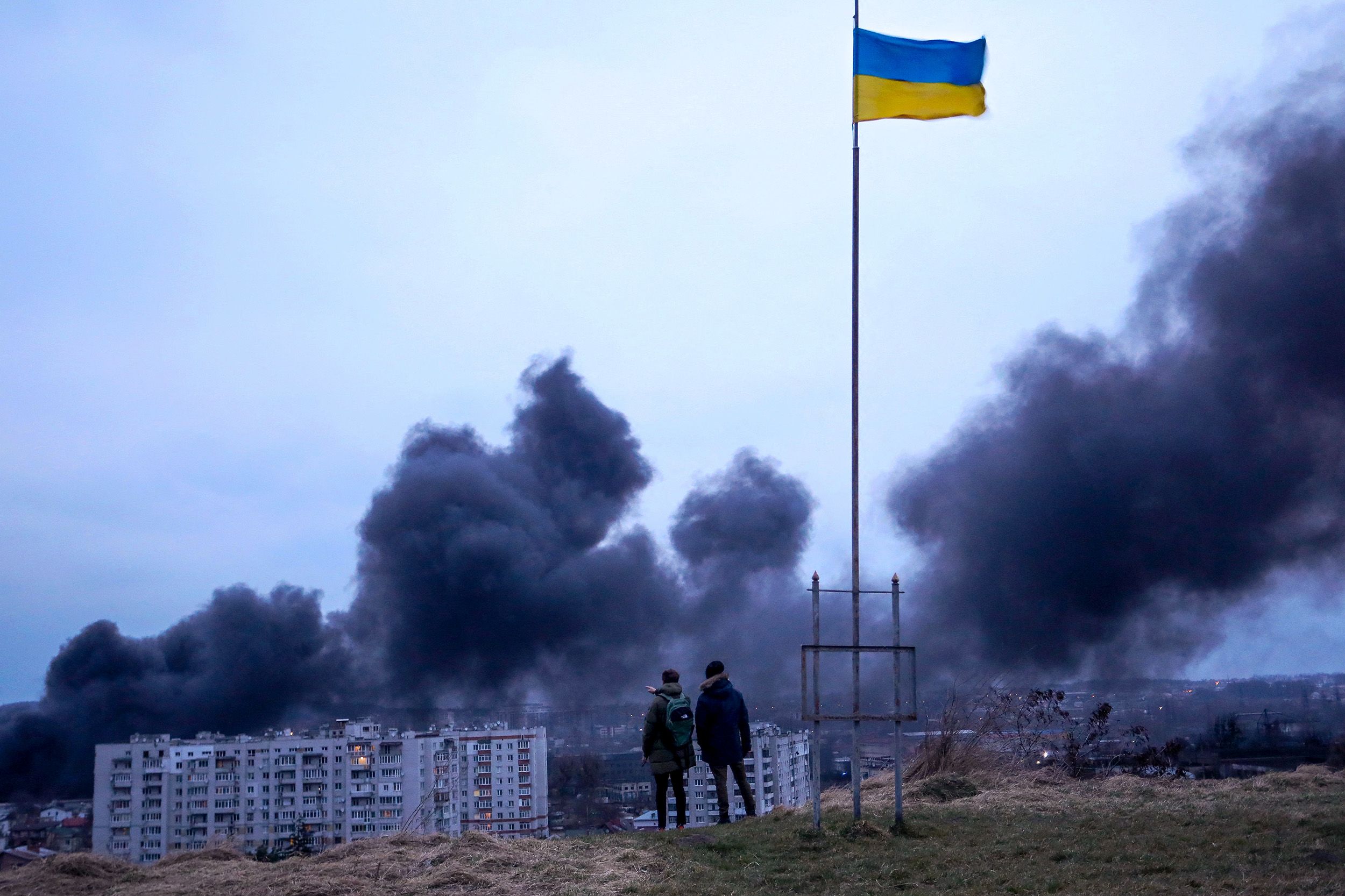 Two people stand on a hill overlooking the destruction of a Ukrainian city with a tall Ukrainian flag flying