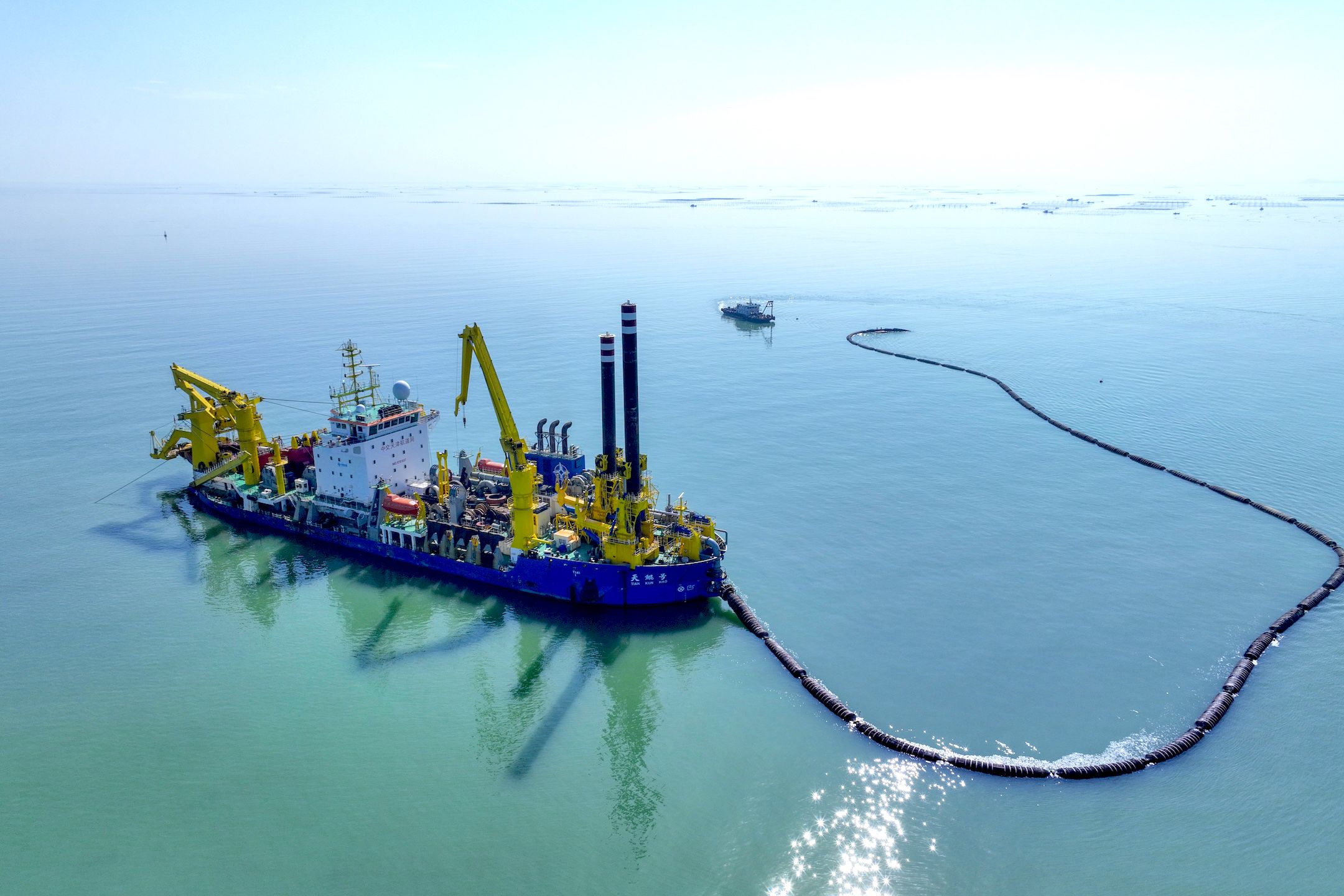 A long pipe connected to a blue and yellow boat with heavy machinery sits in the middle of the ocean