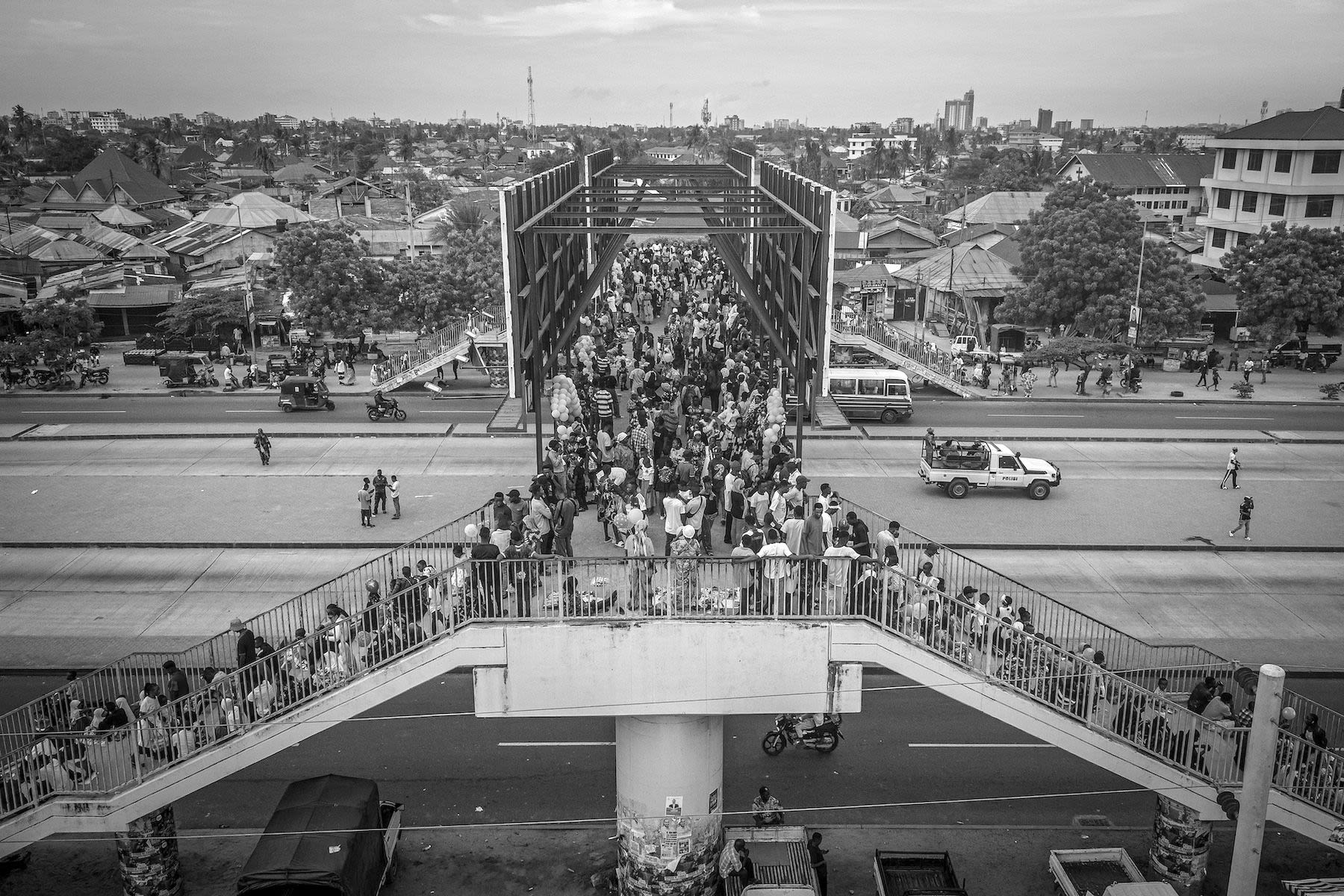 People crowd on a bridge overpassing a street.