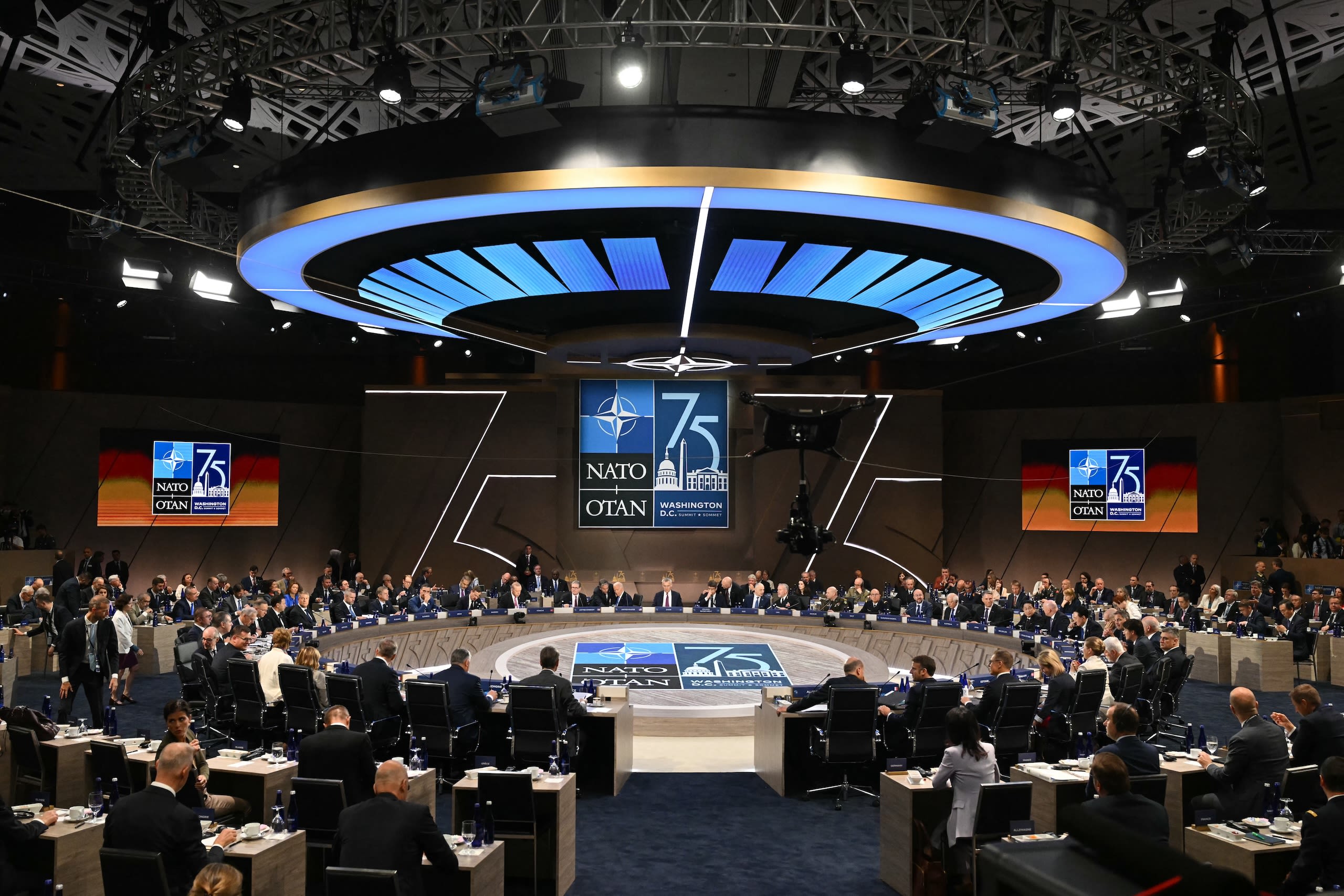 People in suits circle around a large round circular table with the words "NATO" in the middle.